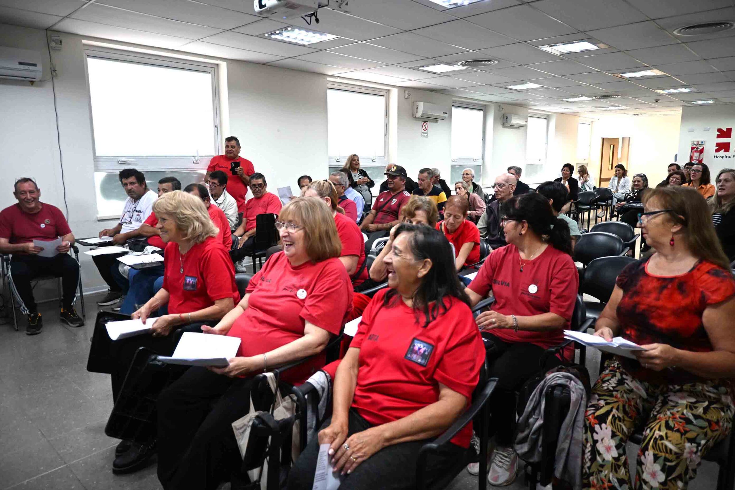 Grupo de hombres y mujeres de diversas edades, pacientes de Parkinson, integrantes del coro 'Voces que inspiran', en un salón del Hospital Rawson. Se los ve animados antes de su presentación por el Día Mundial del Parkinson.