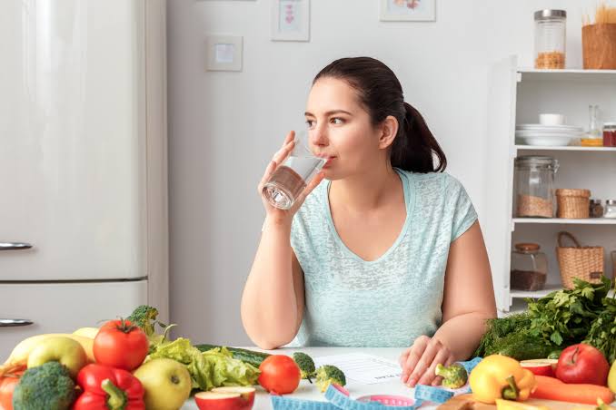 Mujer toma un vaso con agua, mientras también se muestras alimentos con fibra.